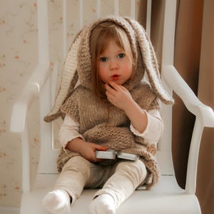 May include: A young child wearing a knitted bunny ear hooded vest, beige pants, and white socks, seated on a white wooden rocking chair. The child is holding a small, silver container. The background features floral wallpaper and a brown curtain.