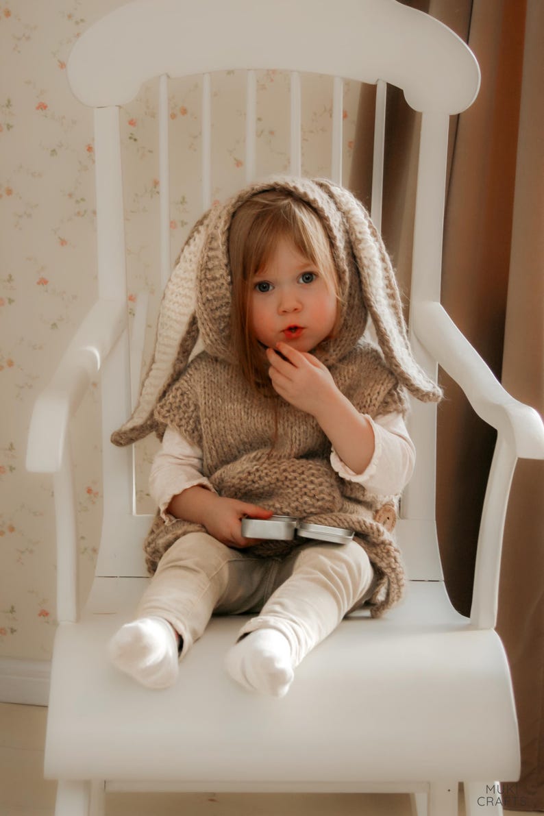 May include: A young child wearing a knitted bunny ear hooded vest, beige pants, and white socks, seated on a white wooden rocking chair. The child is holding a small, silver container. The background features floral wallpaper and a brown curtain.