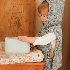 May include: A child's hooded knit vest in gray and white, fastened with a wooden button. The child is wearing a white long-sleeved shirt and tan pants. A light blue cylindrical object sits on a floral patterned cushion.