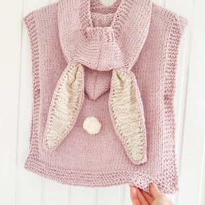 May include: A light pink knitted bunny poncho with a hood and large bunny ears. The ears are beige with pink trim. A small white pom-pom is centered on the front. The poncho is hanging on a white hanger.
