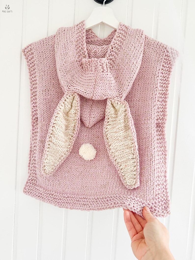 May include: A light pink knitted bunny poncho with a hood and large bunny ears. The ears are beige with pink trim. A small white pom-pom is centered on the front. The poncho is hanging on a white hanger.