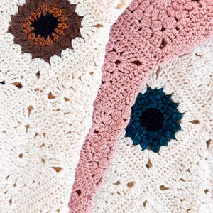 May include: Close-up of a handmade crochet blanket featuring a floral pattern. The blanket has a cream-colored base with brown, teal, and pink floral accents. The flowers have a dark center with a glittery gold outline. The blanket is made of soft yarn.