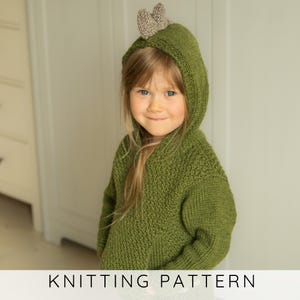 May include: A child wearing a green knitted hooded sweater with a small, brown, heart-shaped detail on top. The sweater has a textured appearance. The text "KNITTING PATTERN" is displayed at the bottom of the image.
