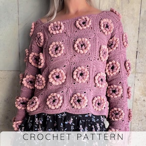 Puede incluir: Un suéter de ganchillo malva con detalles florales en relieve. El suéter presenta un patrón cuadrado con múltiples flores tridimensionales en tonos rosas. Las mangas son largas y el cuello es ancho. El texto "CROCHET PATTERN" está en la parte inferior.
