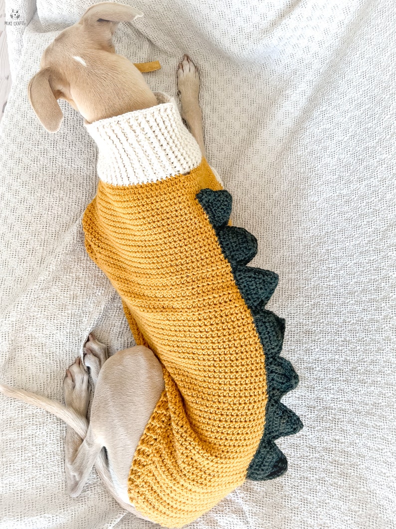 May include: A dog sweater featuring a mustard yellow body, a white ribbed turtleneck collar, and a row of dark green crocheted spikes running down the back. The garment is crafted with a textured stitch pattern and is worn by a light brown dog.