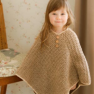 May include: A tan crochet poncho with three wooden buttons at the neckline. The poncho is worn by a young child. The crochet stitch creates a textured appearance. The child is wearing beige pants. The background includes a floral wallpaper and a wooden chair.