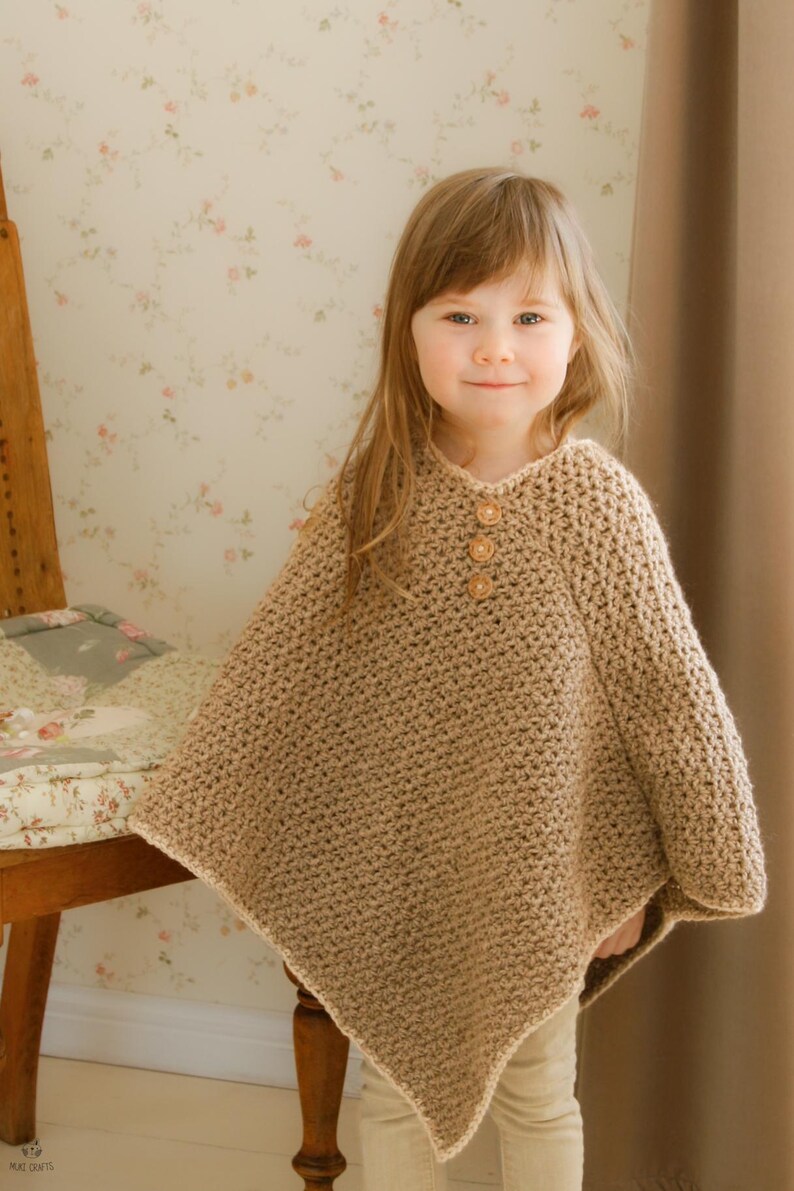 May include: A tan crochet poncho with three wooden buttons at the neckline. The poncho is worn by a young child. The crochet stitch creates a textured appearance. The child is wearing beige pants. The background includes a floral wallpaper and a wooden chair.