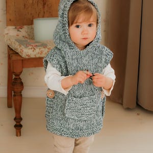 May include: A child wearing a gray and white knitted hooded vest with a front pocket and wooden buttons. The child is also wearing a white long-sleeved shirt and tan pants. The vest is handmade and appears cozy.