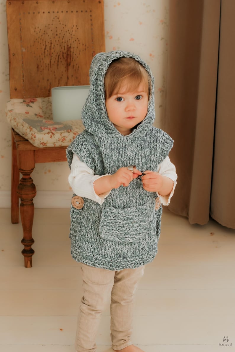 May include: A child wearing a gray and white knitted hooded vest with a front pocket and wooden buttons. The child is also wearing a white long-sleeved shirt and tan pants. The vest is handmade and appears cozy.