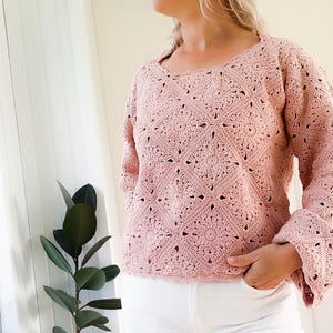 Op de afbeelding: Een lichtroze gehaakte trui met lange mouwen en een vierkante halslijn. De trui heeft een openwerk ontwerp met bloemen en geometrische patronen. De mouwen hebben een uitlopende vorm. De persoon draagt een witte broek.