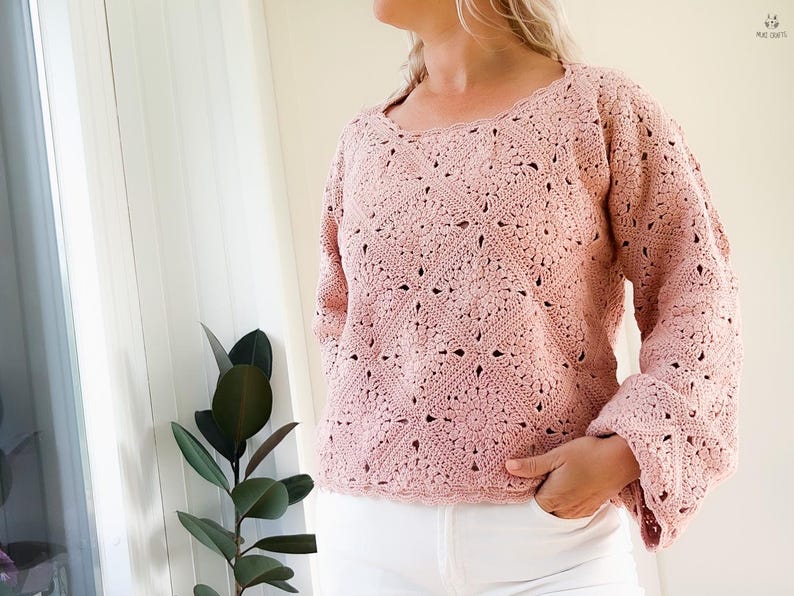 Op de afbeelding: Een lichtroze gehaakte trui met lange mouwen en een vierkante halslijn. De trui heeft een openwerk ontwerp met bloemen en geometrische patronen. De mouwen hebben een uitlopende vorm. De persoon draagt een witte broek.