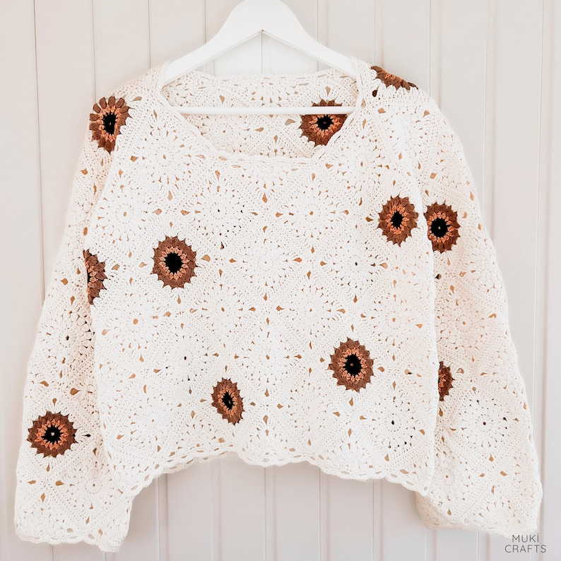 Op de afbeelding: Een witte gehaakte trui met vierkante halslijn, versierd met bruine en zwarte zonnebloemmotieven. Het kledingstuk met lange mouwen wordt gepresenteerd op een witte hanger tegen een witte achtergrond. Het haakpatroon is ingewikkeld en gedetailleerd.