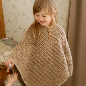 May include: A tan crochet poncho with three wooden buttons at the neckline. The poncho is worn by a young child. The background includes a floral wallpaper and a wooden bed frame. The poncho is a cozy and stylish garment.