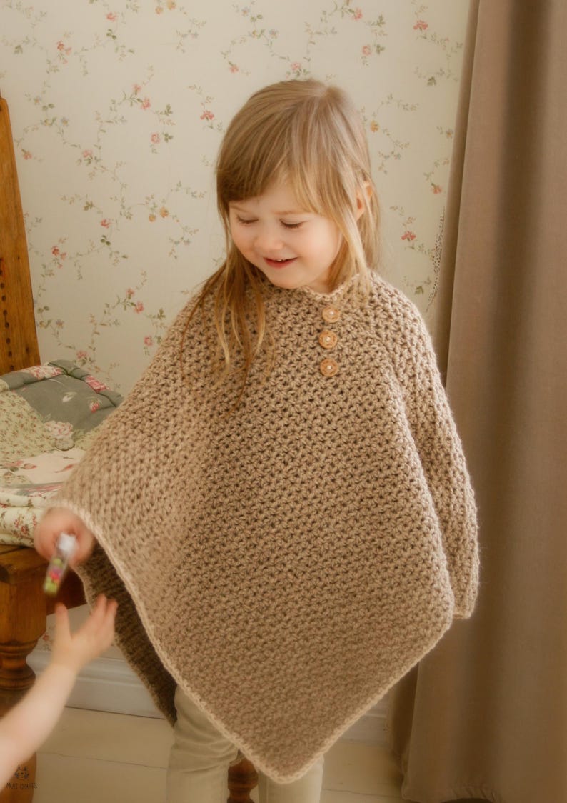May include: A tan crochet poncho with three wooden buttons at the neckline. The poncho is worn by a young child. The background includes a floral wallpaper and a wooden bed frame. The poncho is a cozy and stylish garment.