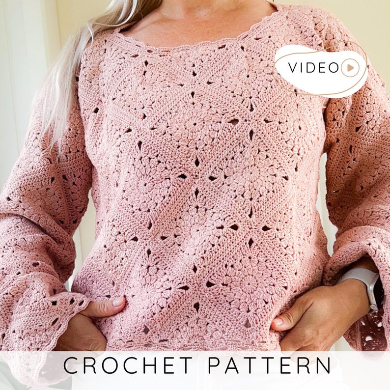 Op de afbeelding: Een lichtroze gehaakte trui met een bloemenpatroon. De top met lange mouwen heeft een geschulpte halslijn en uitlopende mouwen. De tekst "CROCHET PATTERN" wordt onderaan de afbeelding weergegeven. Een video-icoon staat rechtsboven.