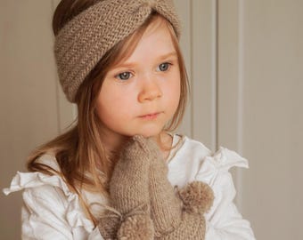 May include: A light brown knitted headband with a central twist detail and matching knitted mittens featuring small pom-poms. The set is displayed with the text KNITTING PATTERN visible at the bottom of the image, indicating a guide for creation.