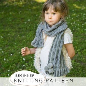 Puede incluir: Una bufanda de punto gris con textura acanalada, extremos con volantes y tres pequeños pompones unidos a un lado. La bufanda se lleva sobre una camiseta blanca y un chaleco de piel sintética color crema. El texto en la imagen dice: BEGINNER KNITTING PATTERN.