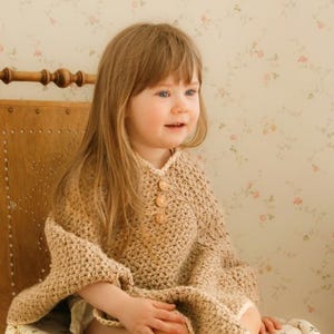 May include: A tan crochet poncho with three wooden buttons. The poncho is worn by a young child with long blonde hair, seated on a wooden chair with a floral cushion. The background features a floral wallpaper.