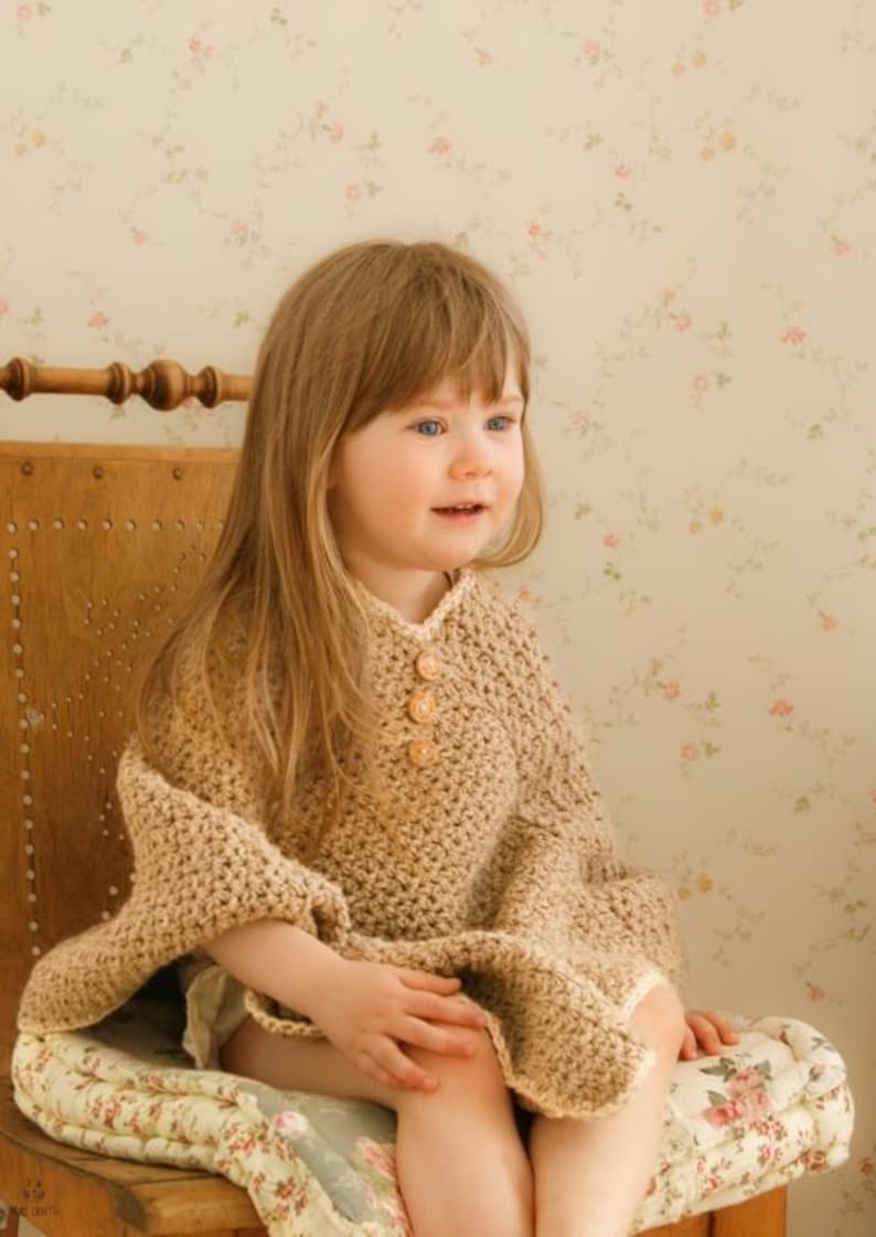 May include: A tan crochet poncho with three wooden buttons. The poncho is worn by a young child with long blonde hair, seated on a wooden chair with a floral cushion. The background features a floral wallpaper.