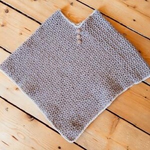 May include: A gray knitted poncho with a V-neck and three wooden buttons. The poncho has a textured appearance and a white trim along the edges. The garment is displayed on a wooden surface.