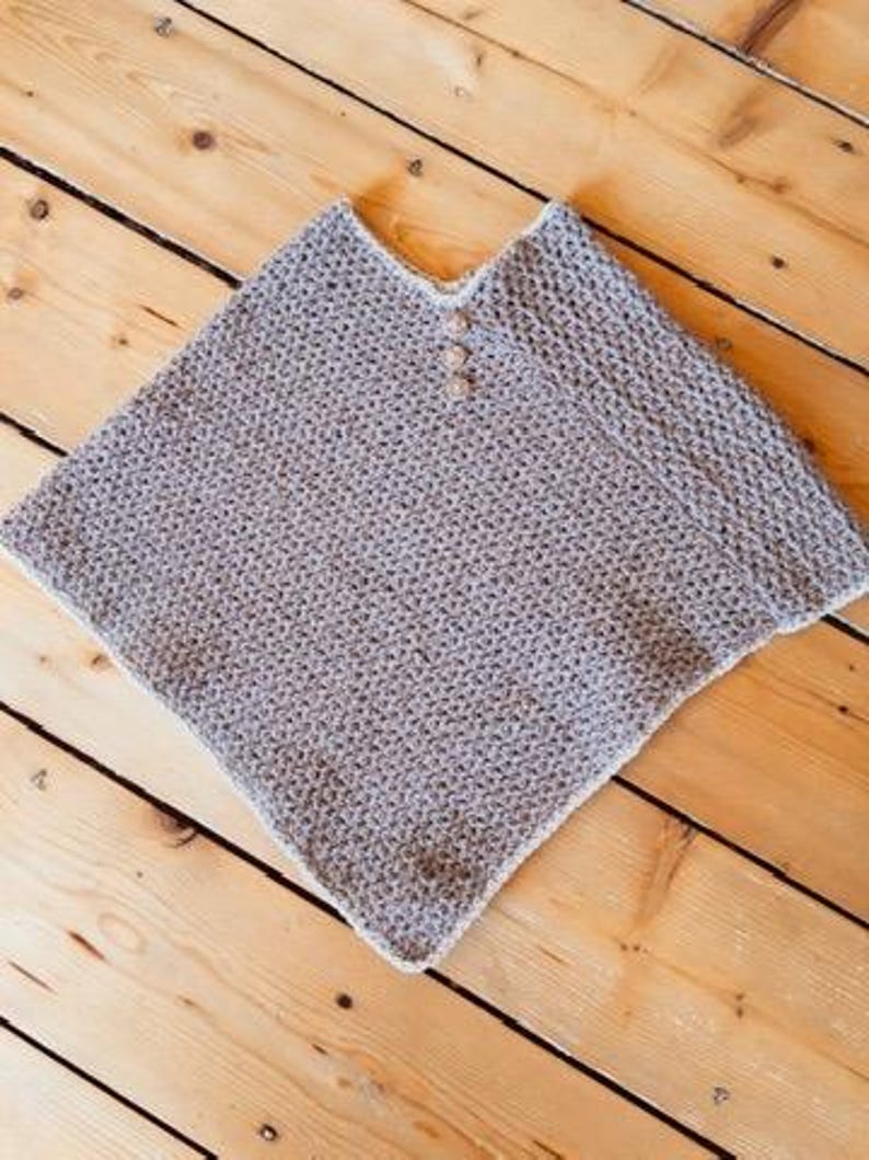 May include: A gray knitted poncho with a V-neck and three wooden buttons. The poncho has a textured appearance and a white trim along the edges. The garment is displayed on a wooden surface.