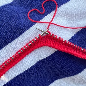 May include: A close-up of red knitting in progress, with two silver needles holding the stitches. The red yarn forms a heart shape above the knitting. The background is a blue and white striped towel.
