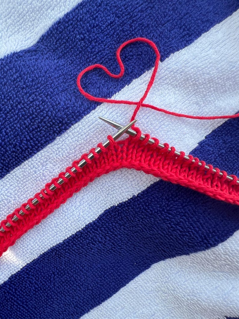 May include: A close-up of red knitting in progress, with two silver needles holding the stitches. The red yarn forms a heart shape above the knitting. The background is a blue and white striped towel.