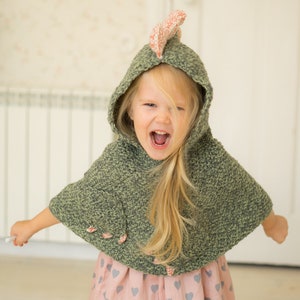 Peut inclure: Un enfant porte une cape &agrave; capuche en crochet vert avec un accent rose et blanc sur la capuche. La cape a des boutons en forme de fleurs roses. L'enfant porte &eacute;galement une robe rose avec des motifs de c&oelig;urs gris.