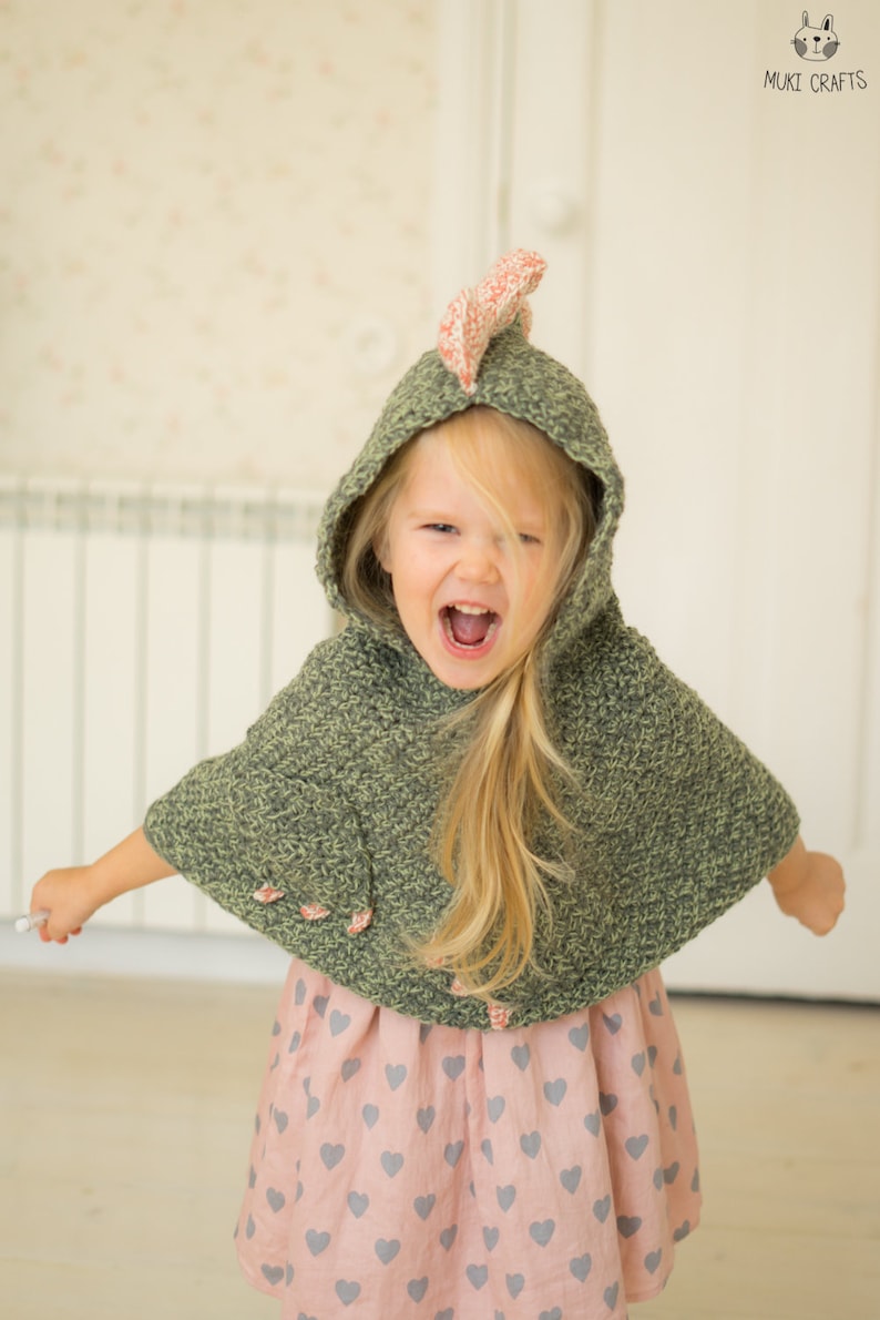 Peut inclure: Un enfant porte une cape &agrave; capuche en crochet vert avec un accent rose et blanc sur la capuche. La cape a des boutons en forme de fleurs roses. L'enfant porte &eacute;galement une robe rose avec des motifs de c&oelig;urs gris.