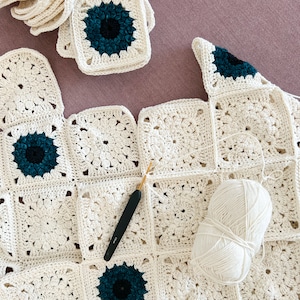 May include: A work in progress, a crochet blanket with cream-colored squares and teal and black floral centers. A crochet hook and a ball of white yarn are on the blanket. Several finished squares are stacked nearby.