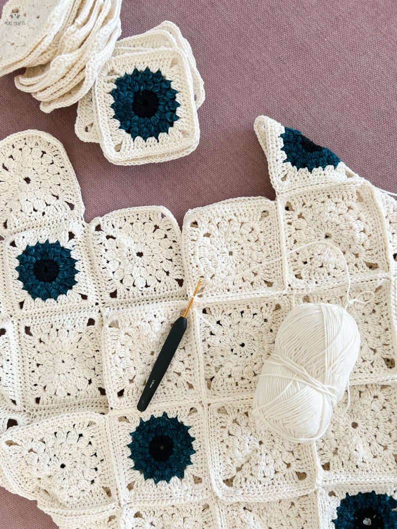 May include: A work in progress, a crochet blanket with cream-colored squares and teal and black floral centers. A crochet hook and a ball of white yarn are on the blanket. Several finished squares are stacked nearby.
