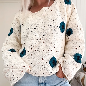 Close‑up of a cream, granny‑square crochet sweater worked on the diagonal and scattered with teal, sunflower‑like motifs; the relaxed, scallop‑neck pullover is modelled with the sleeves slightly scrunched