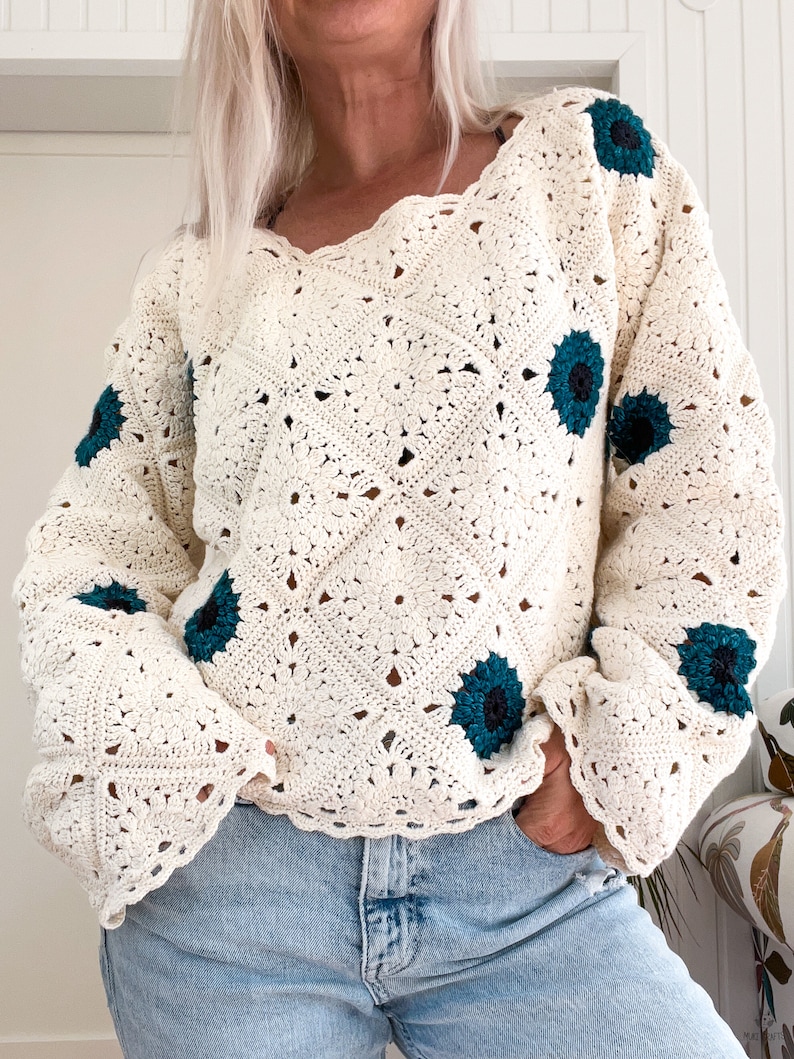 Close‑up of a cream, granny‑square crochet sweater worked on the diagonal and scattered with teal, sunflower‑like motifs; the relaxed, scallop‑neck pullover is modelled with the sleeves slightly scrunched