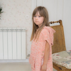 May include: A child wearing a coral and white knit hooded poncho with wooden buttons. The poncho has a pocket and is paired with light pink leggings. The background includes a floral wallpaper, a wooden chair, and a radiator.