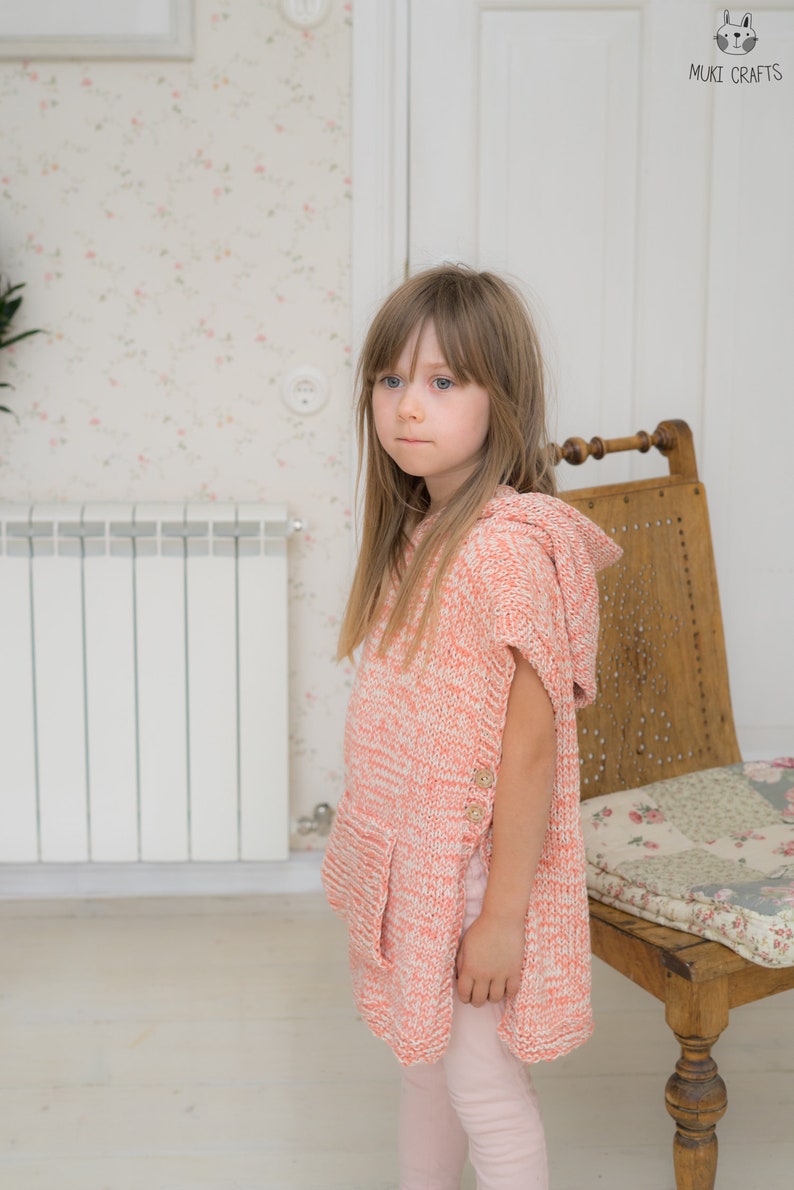May include: A child wearing a coral and white knit hooded poncho with wooden buttons. The poncho has a pocket and is paired with light pink leggings. The background includes a floral wallpaper, a wooden chair, and a radiator.