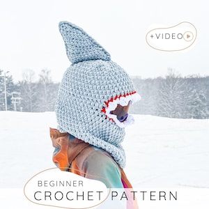 Può includere: Un cappello da squalo all'uncinetto grigio chiaro con una pinna appuntita e un bordo rosso e bianco attorno all'apertura della bocca. Il cappello fa parte di un modello all'uncinetto per principianti. Lo sfondo è un paesaggio innevato.