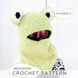 Könnte beinhalten: Eine hellgrüne gehäkelte Frosch-Balaclava mit weißen und schwarzen Akzenten. Die Balaclava hat eine Gesichtsöffnung für Skibrillen. Der Text "BEGINNER CROCHET PATTERN" wird am unteren Rand des Bildes angezeigt.