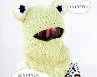 Op de afbeelding: Een lichtgroene gehaakte kikkerbalaclava met witte en zwarte accenten. De balaclava heeft een gezichtsopening voor een skibril. De tekst "BEGINNER CROCHET PATTERN" wordt onderaan de afbeelding weergegeven.