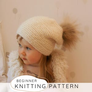 May include: A cream-colored knitted beanie hat featuring a textured garter stitch pattern and a large, fluffy, light brown faux fur pom-pom attached to the top. The image includes the text BEGINNER KNITTING PATTERN at the bottom of the frame.