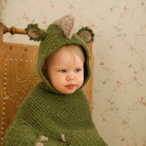 Peut inclure: Un poncho vert pour b&eacute;b&eacute;, crochet&eacute;, avec une capuche en forme de dinosaure. La capuche pr&eacute;sente une cr&ecirc;te et des oreilles beiges. Le poncho est port&eacute; par un b&eacute;b&eacute; assis sur une chaise en bois.