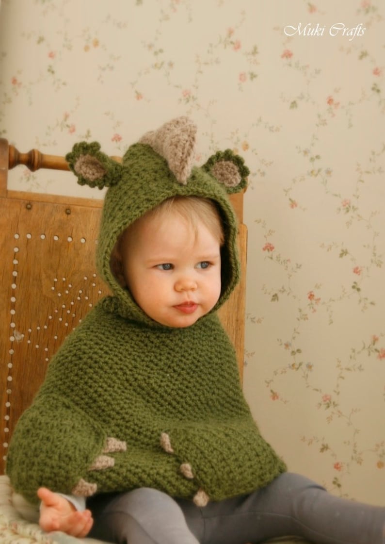Peut inclure: Un poncho vert pour b&eacute;b&eacute;, crochet&eacute;, avec une capuche en forme de dinosaure. La capuche pr&eacute;sente une cr&ecirc;te et des oreilles beiges. Le poncho est port&eacute; par un b&eacute;b&eacute; assis sur une chaise en bois.