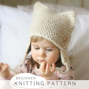 Puede incluir: Un gorro beige tejido con orejeras y dos botones de madera. El gorro lo lleva un niño. El texto "BEGINNER KNITTING PATTERN" se muestra en un óvalo blanco en la parte inferior de la imagen.