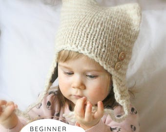 Könnte beinhalten: Eine gestrickte beige Mütze mit Ohrenklappen und zwei Holzknöpfen. Die Mütze wird von einem Kind getragen. Der Text "BEGINNER KNITTING PATTERN" wird in einem weißen Oval am unteren Bildrand angezeigt.
