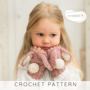 Op de afbeelding: Een paar gehaakte vingerloze handschoenen in oudroze met witte pompons en konijnenoren. De handschoenen hebben een zichtbaar steekpatroon en bevatten de tekst CROCHET PATTERN onderaan en +VIDEO rechtsboven in de afbeelding.