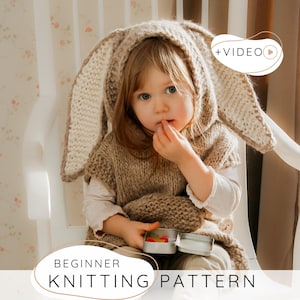 Op de afbeelding: Een kind draagt een gebreide konijnenorenkap en vest, zittend op een witte stoel. De kap en het vest zijn beige. De afbeelding bevat de tekst "BEGINNER KNITTING PATTERN". Een video-icoon staat rechtsboven.