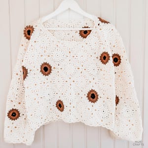 May include: A white, square-neck crochet sweater with brown and black sunflower appliqu&eacute;s. The long-sleeved top is displayed on a white hanger against a white background. The crochet design features a repeating pattern.