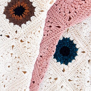 Op de afbeelding: Close-up van een handgemaakte gehaakte deken met een bloemenpatroon. De deken heeft een cr&egrave;mekleurige basis met bruine, blauwgroene en roze bloemaccenten. De haaksteken zijn zichtbaar en cre&euml;ren een textuur.