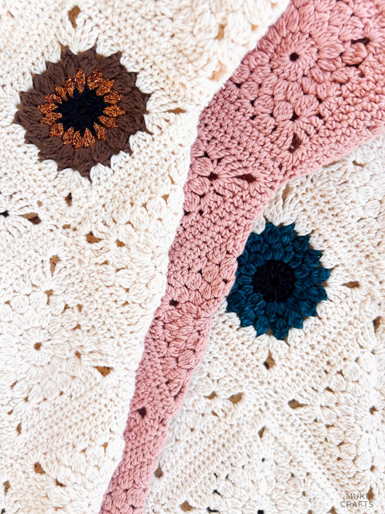 Op de afbeelding: Close-up van een handgemaakte gehaakte deken met een bloemenpatroon. De deken heeft een cr&egrave;mekleurige basis met bruine, blauwgroene en roze bloemaccenten. De haaksteken zijn zichtbaar en cre&euml;ren een textuur.