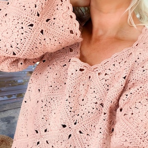 A woman wears a dusty‑pink granny‑square crochet pullover pieced from lacy flower‑motif squares; the relaxed drop‑shoulder sweater has wide sleeves casually pushed up and a scalloped neckline and hem