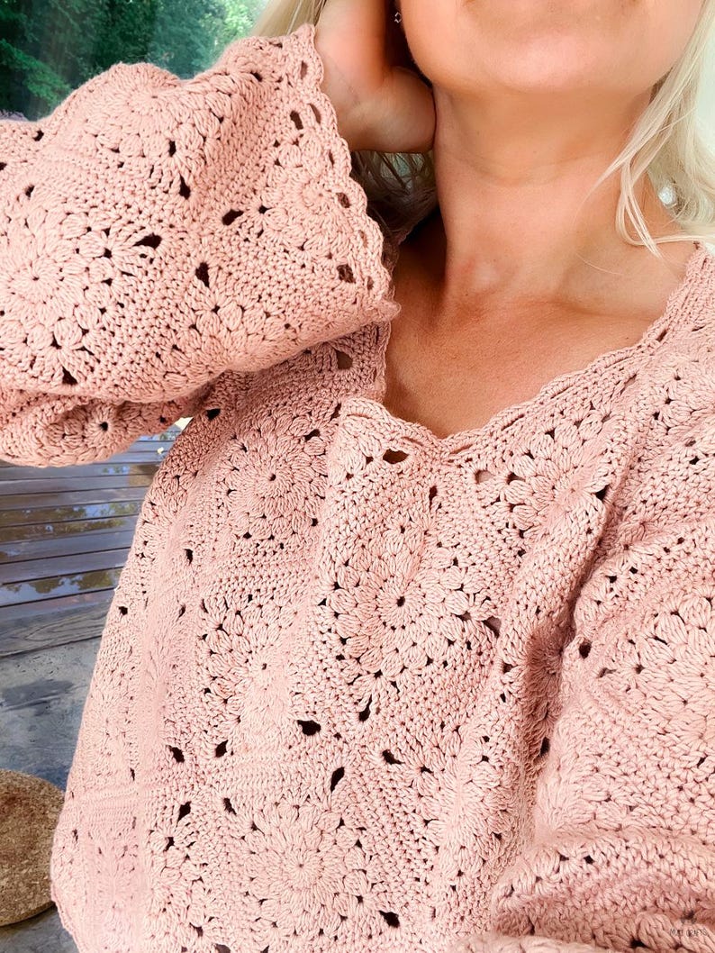 A woman wears a dusty‑pink granny‑square crochet pullover pieced from lacy flower‑motif squares; the relaxed drop‑shoulder sweater has wide sleeves casually pushed up and a scalloped neckline and hem